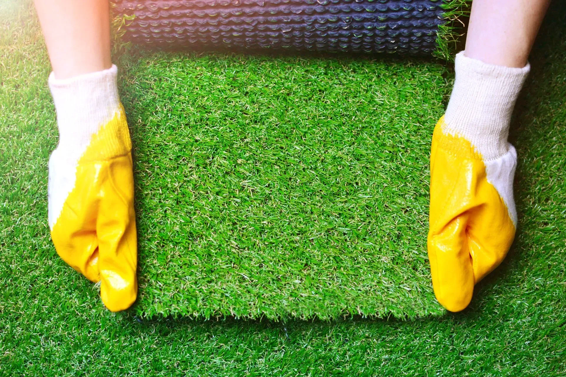 A person wearing yellow gloves is rolling a synthetic grass mat.