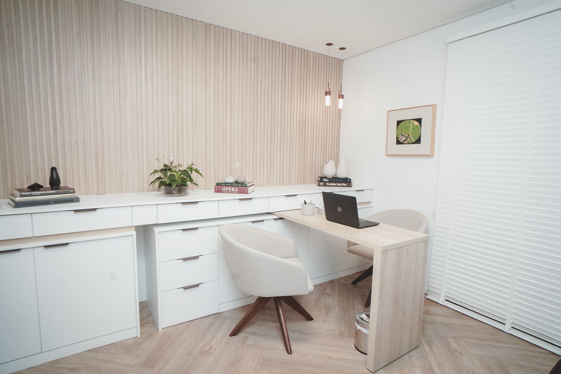 Escritório em casa bem iluminado com mesa, cadeira, armários, persianas e um vaso de plantas brancos.