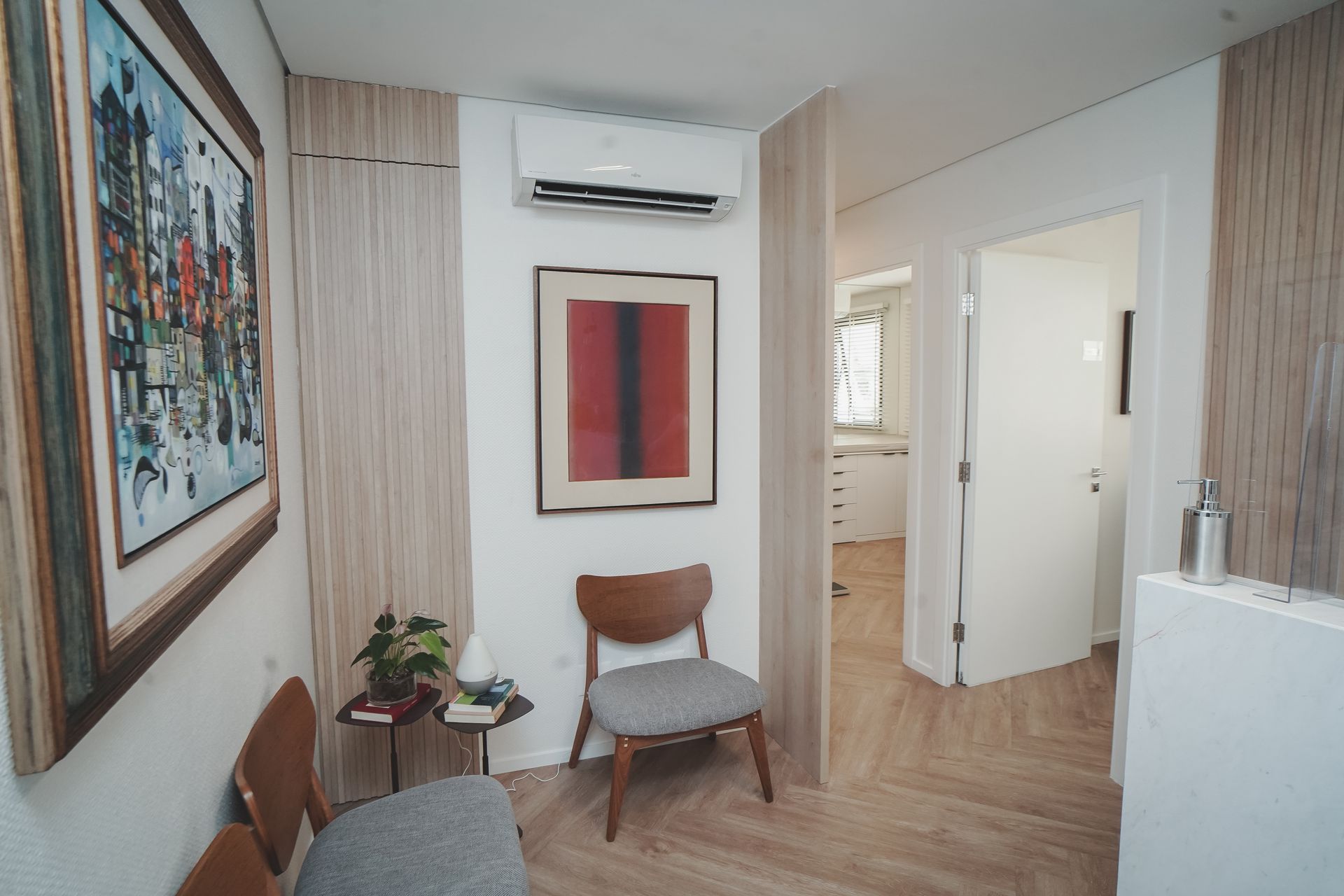 Quarto pequeno e moderno com cadeiras, arte abstrata na parede, ar condicionado e uma porta aberta para um hall iluminado.