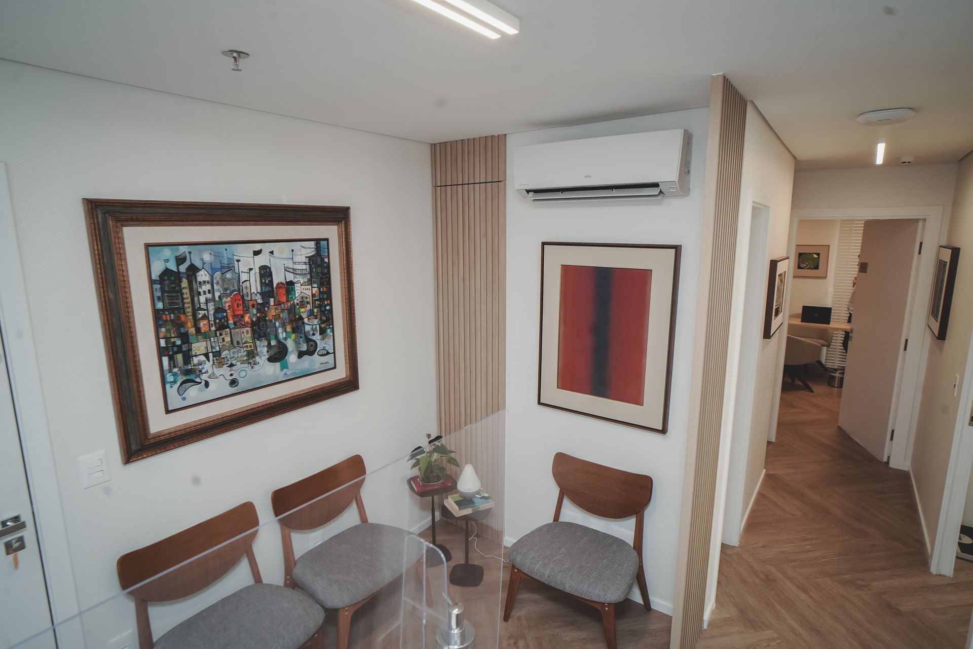 Área de espera moderna com cadeiras, mesa de vidro, obras de arte emolduradas e ar condicionado em um corredor.