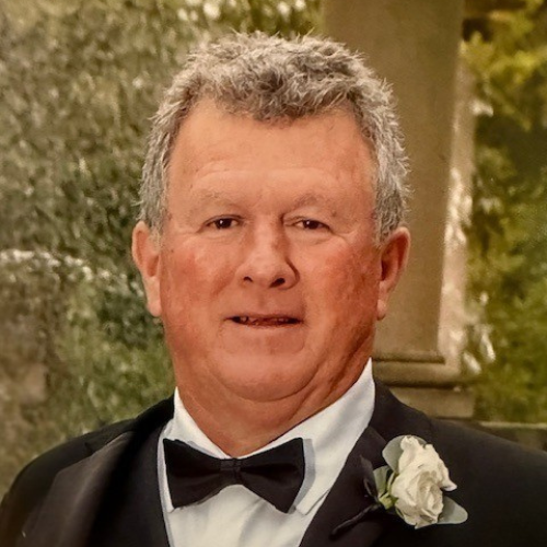 Portrait of a man in a tuxedo with a black bow tie and white boutonniere, standing outdoors.