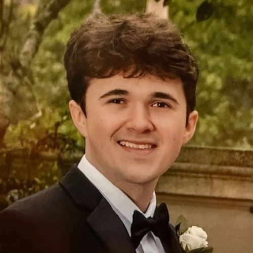 Young man in a black tuxedo with bow tie, smiling in a formal portrait with a soft floral background.