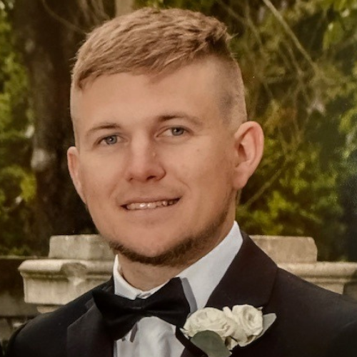 Man in a black tuxedo with white boutonniere, smiling outdoors against a blurred greenery background.
