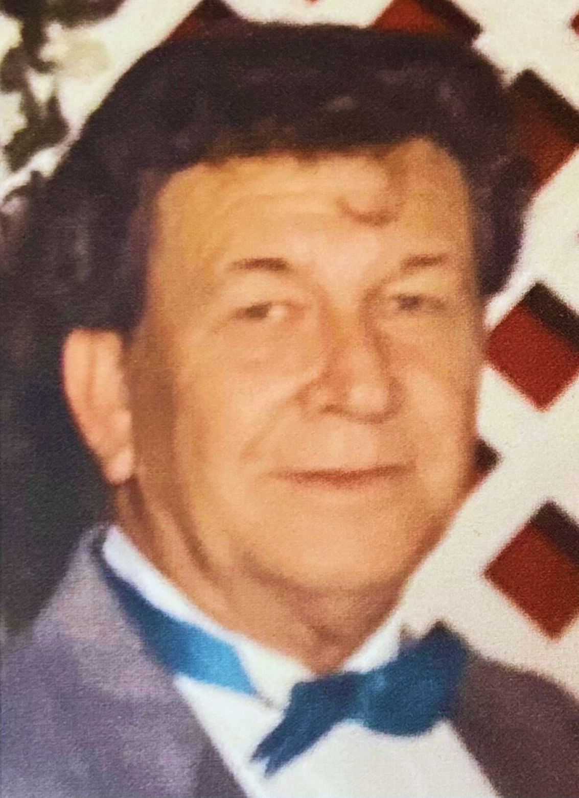 Man in a tuxedo with a blue bow tie, smiling against a red-and-white patterned background