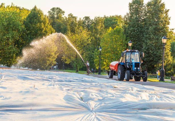 A worker sprays green hydroseed onto a sloped hillside reinforced with erosion control blankets and straw wattles.
