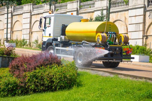 A man sprays green hydroseed onto bare soil from a hose. A large tank trailer sits in the background near a house and trees.