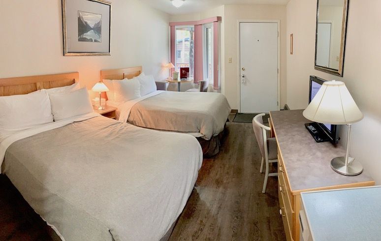 Hotel room with two beds, desk, and a TV. Pale walls and flooring.