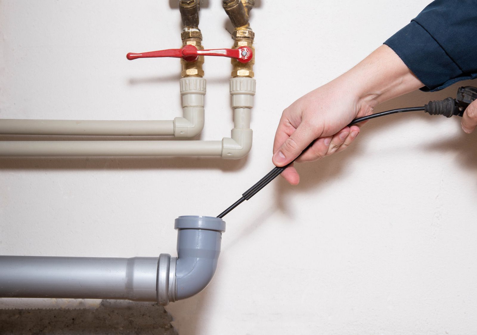 Person using a drain snake to unclog a gray PVC drain pipe near a wall with plumbing.