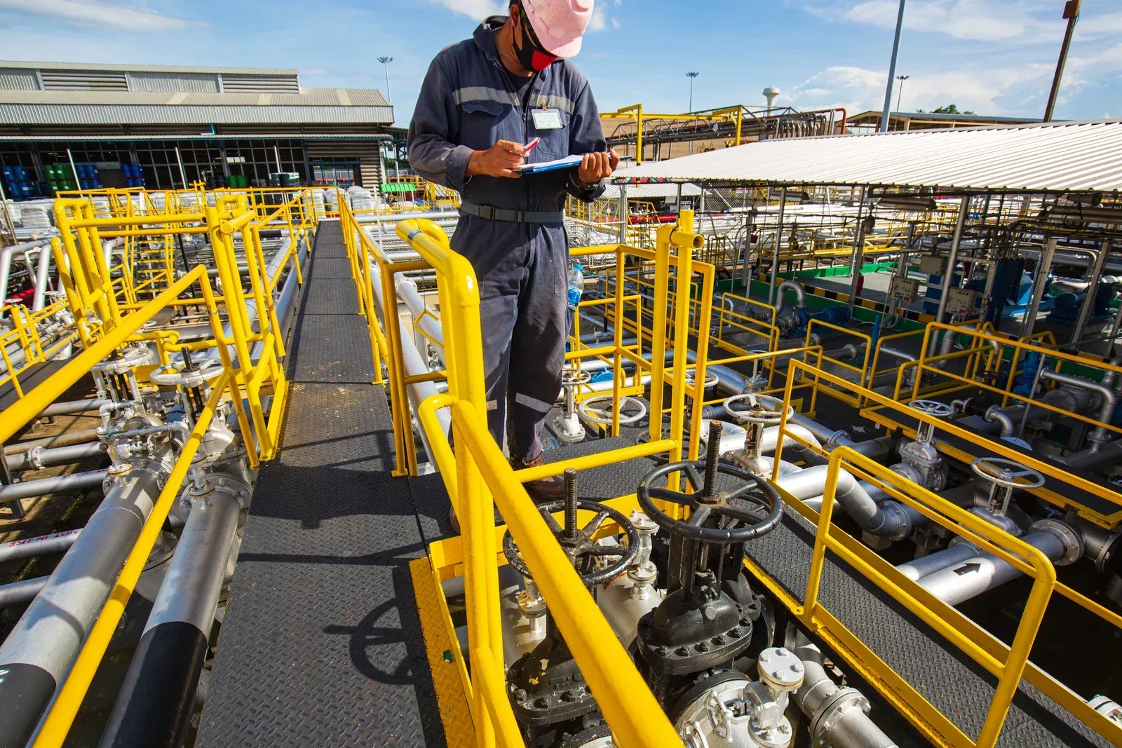 Person in a jumpsuit and hard hat inspects equipment at an industrial site, walking along a yellow-railed walkway, holding a clipboard.