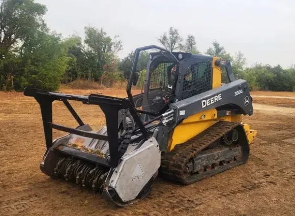 A yellow and gray tractor is sitting in a dirt field. Yellow John Deere mulcher on tracks, in a field, ready for use.