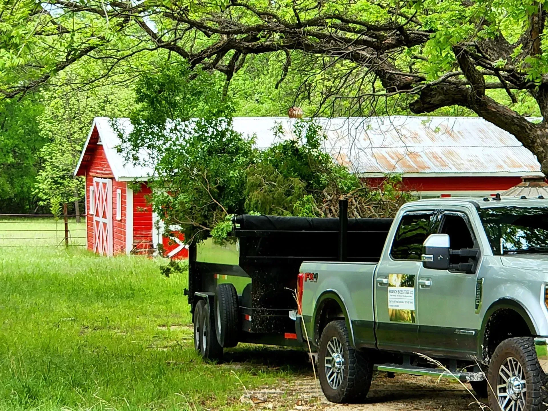 Silver pickup truck with trailer parked on grass near a red barn under leafy trees Silver pickup truck with trailer parked on grass near a red barn under leafy trees
