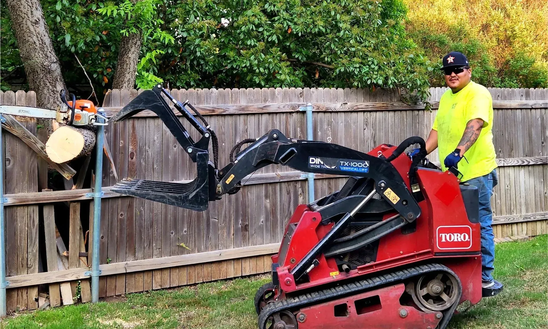 Man operating a red Toro mini skid steer with forks, lifting a tree trunk near a wooden fence.