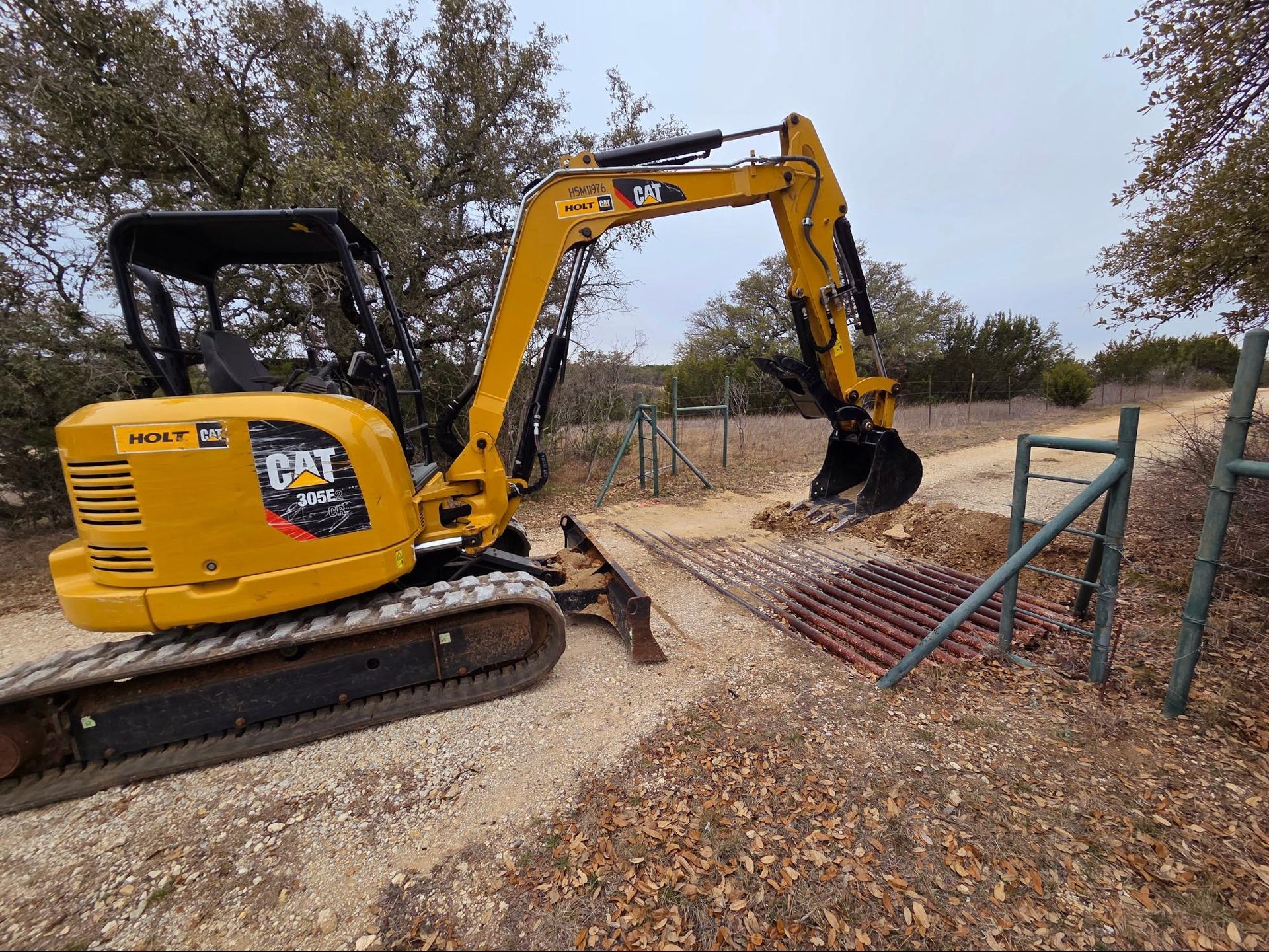 Yellow mini excavator clearing debris beside a metal gate on a gravel driveway.