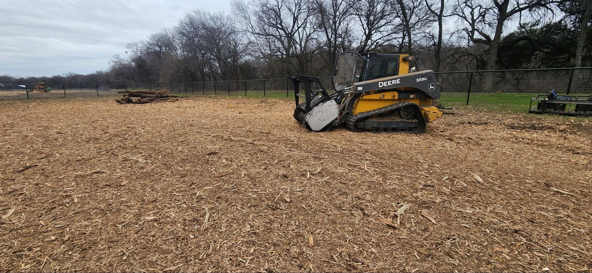 A close up of a pile of brown bark. A yellow skid steer tractor working on a field of wood chips near trees and a fence.