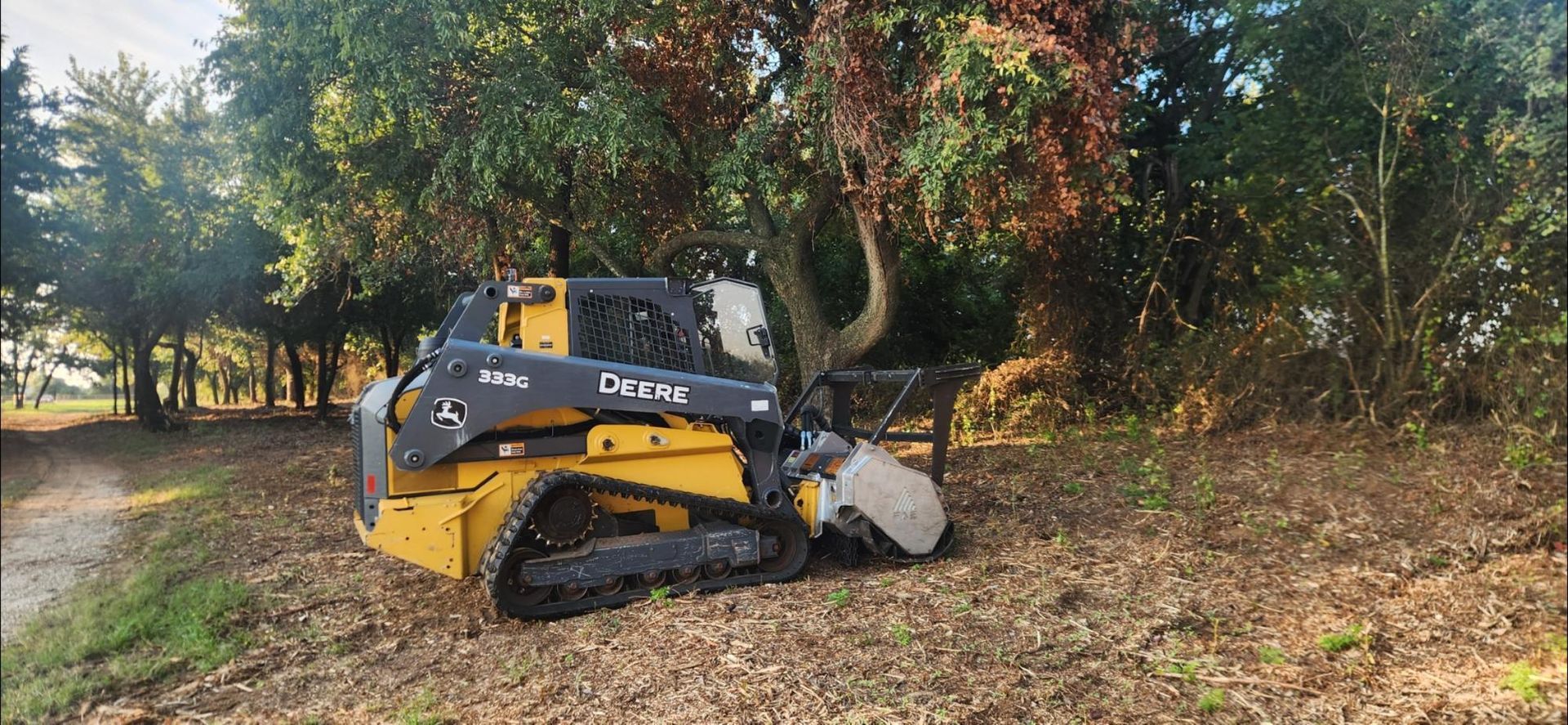 A yellow Deere D-series compact track loader with a brush cutter attachment clearing brush near trees.