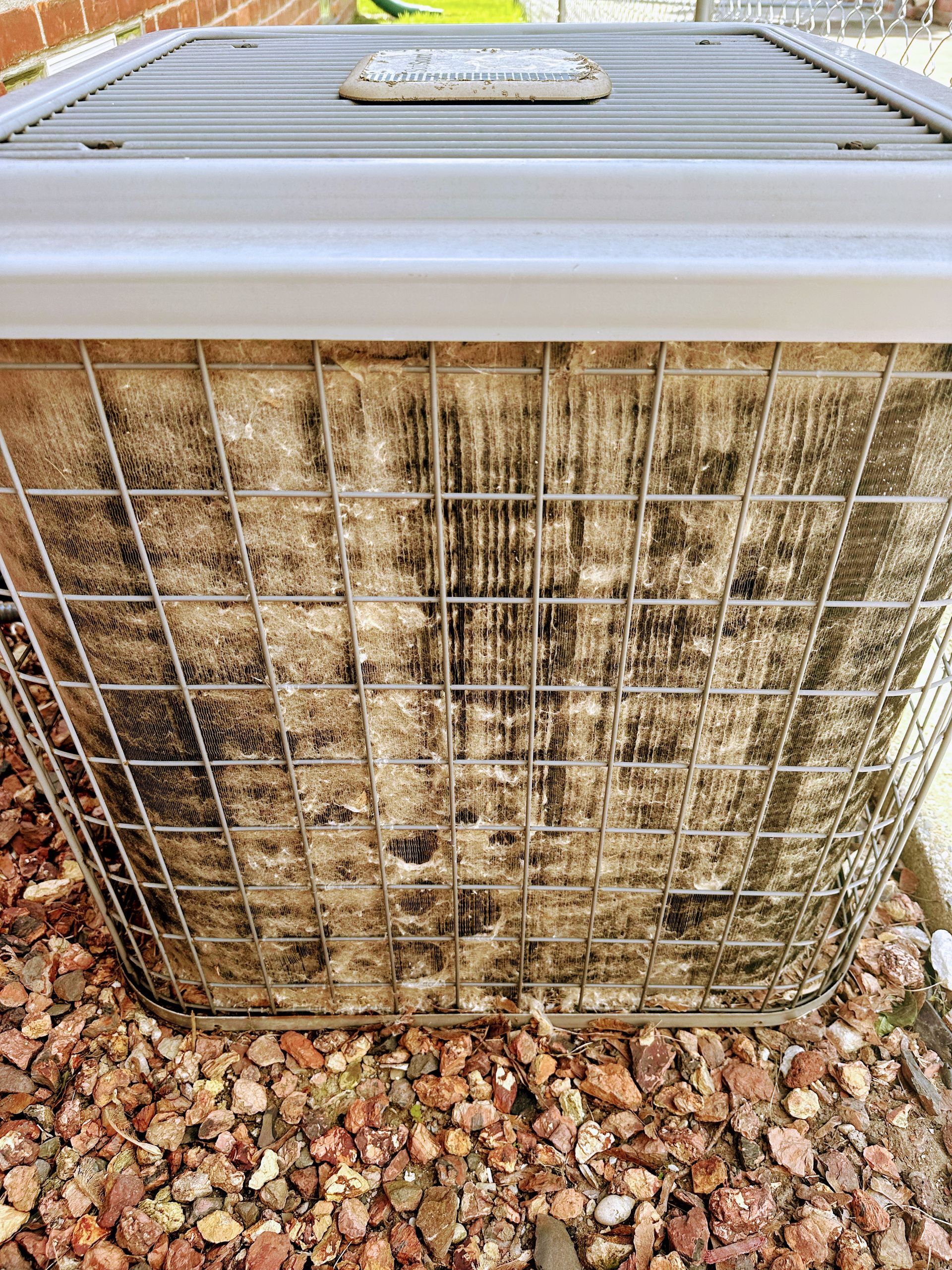 Dirty air conditioner unit with metal cage and black residue on the fins, sitting on gravel.