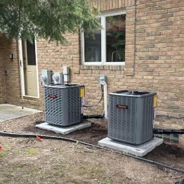 Two gray HVAC units on concrete pads beside a brick house with a window and doorway Two gray HVAC units on concrete pads beside a brick house with a window and doorway