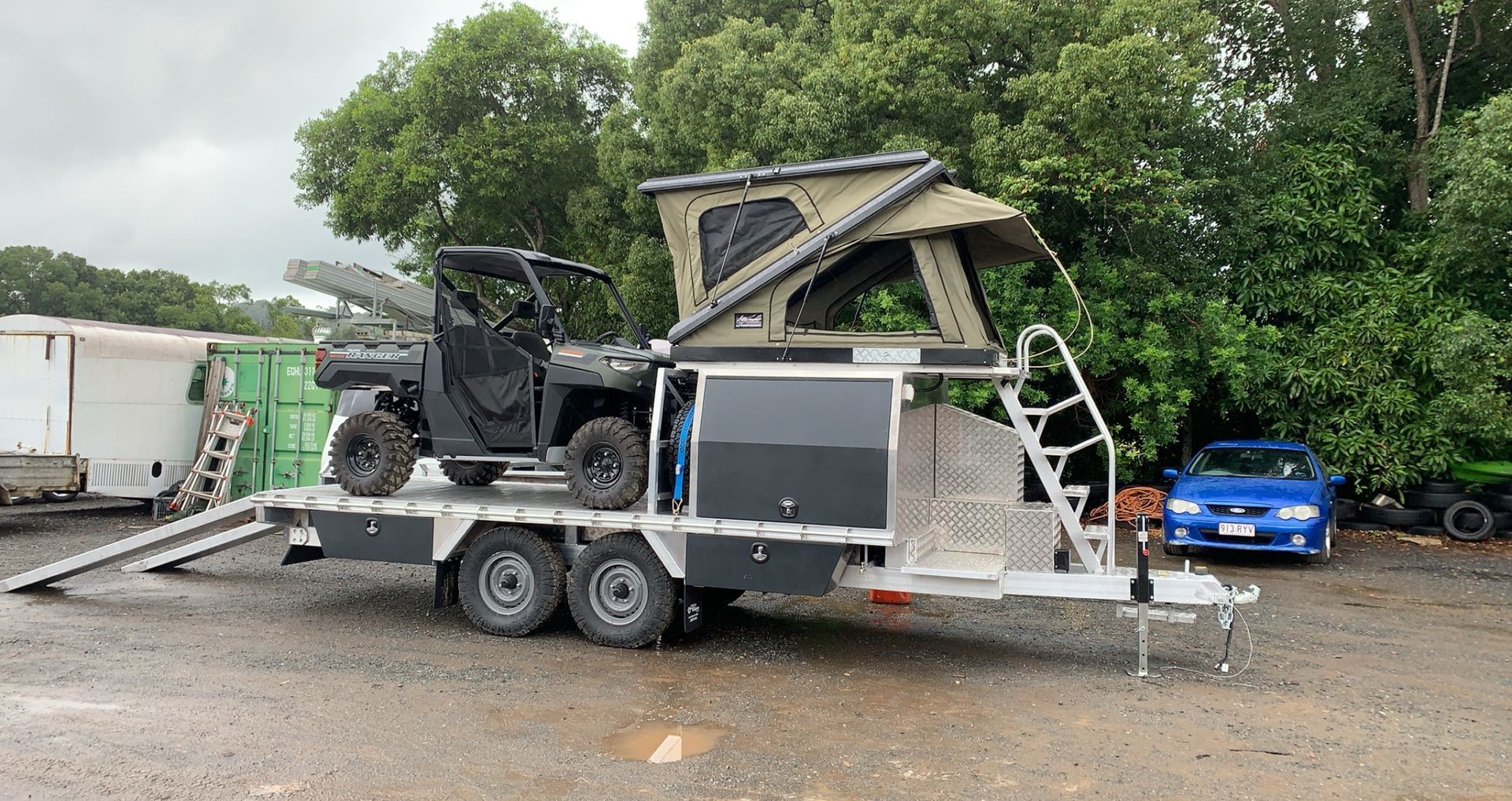 A tandem-axle trailer carrying an ATV and a rooftop tent, parked on a gravel lot next to trees and other vehicles — Rod's Engineering & Lifetime Trailers in Bli Bli, QLD