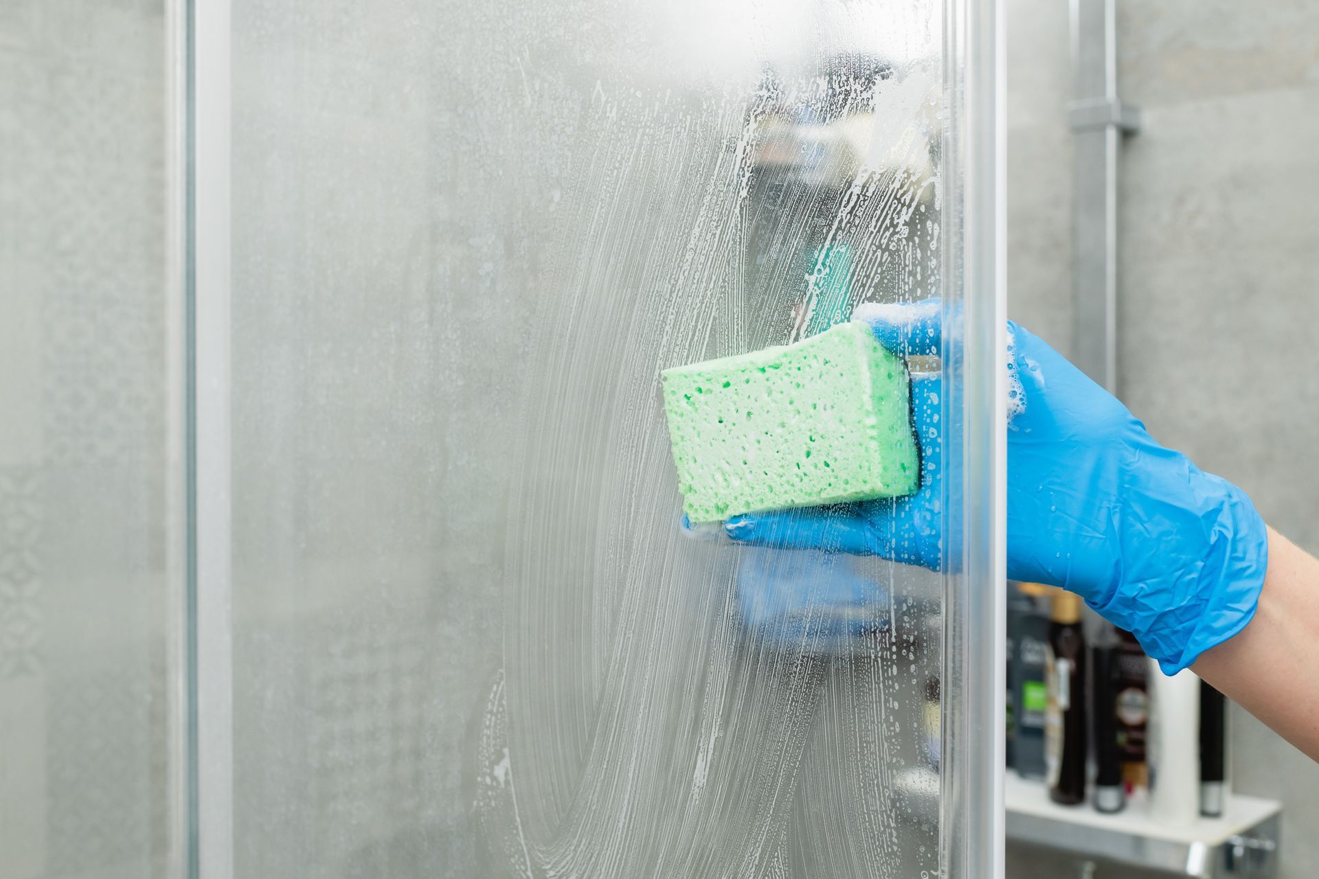 Gloved hand scrubbing a soapy glass shower door with a green sponge.