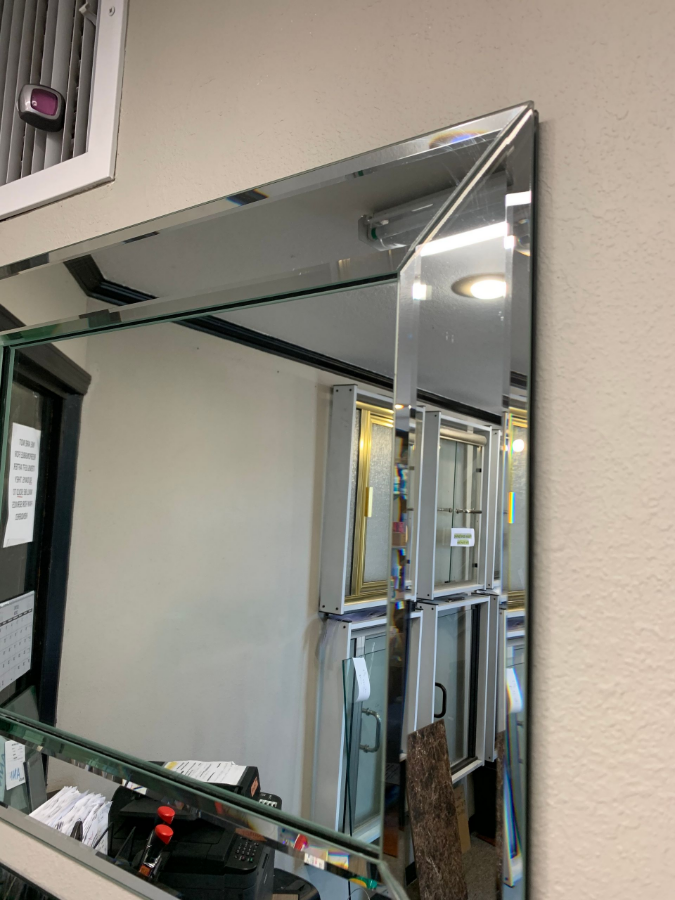 Large rectangular beveled mirror on a cream wall reflecting a cluttered room with windows.