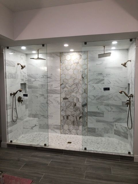 Three-stall shower with glass doors and marble tile. Each stall has a showerhead and handheld wand with bronze fixtures.