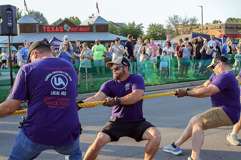 Men in purple shirts tugging rope in a tug-of-war competition, outdoors with a crowd watching near a Texas Roadhouse.