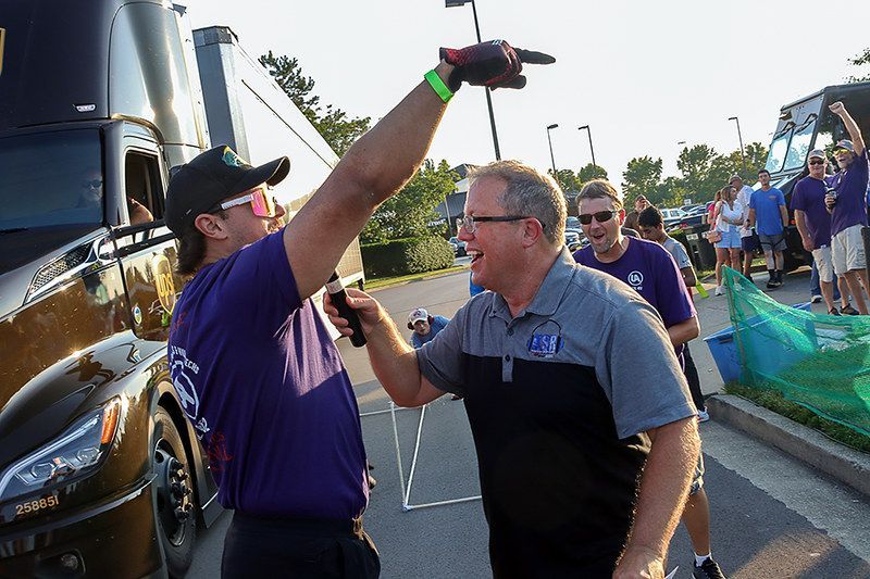 Two men celebrating, pointing. One wears a hat, shades, glove. Other has a logo shirt. Outdoors by vehicles.
