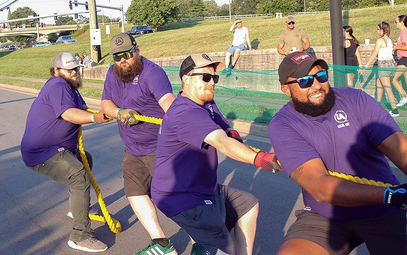 Men in purple shirts and sunglasses tugging a rope in a tug-of-war. Sunny outdoor setting.