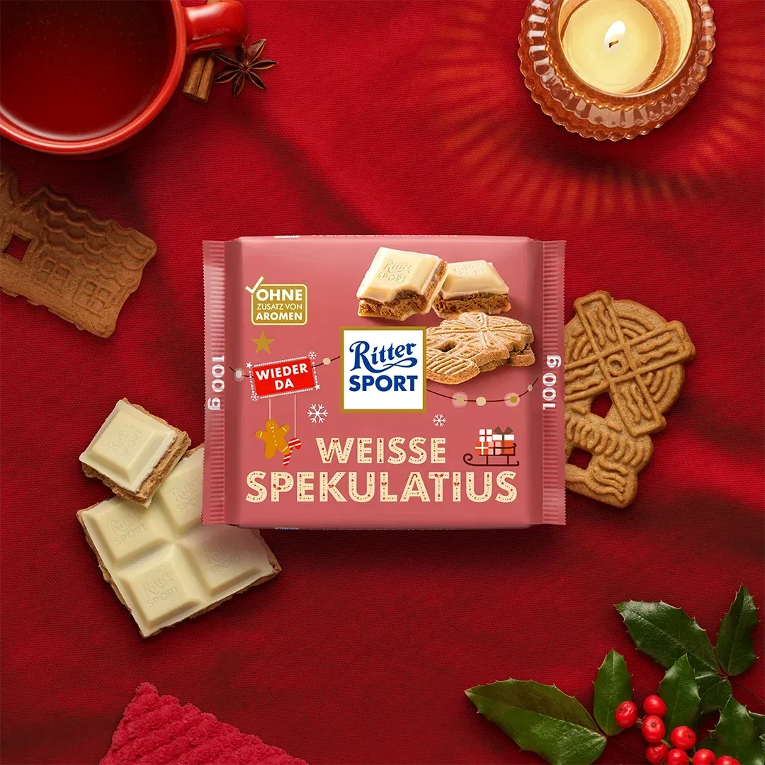Gold Rais Sport Nougat Kugeln Verpackung mit Weihnachtsbaummotiv auf beigem Hintergrund, umgeben von Ornamenten - Bild erstellt mit KI.
