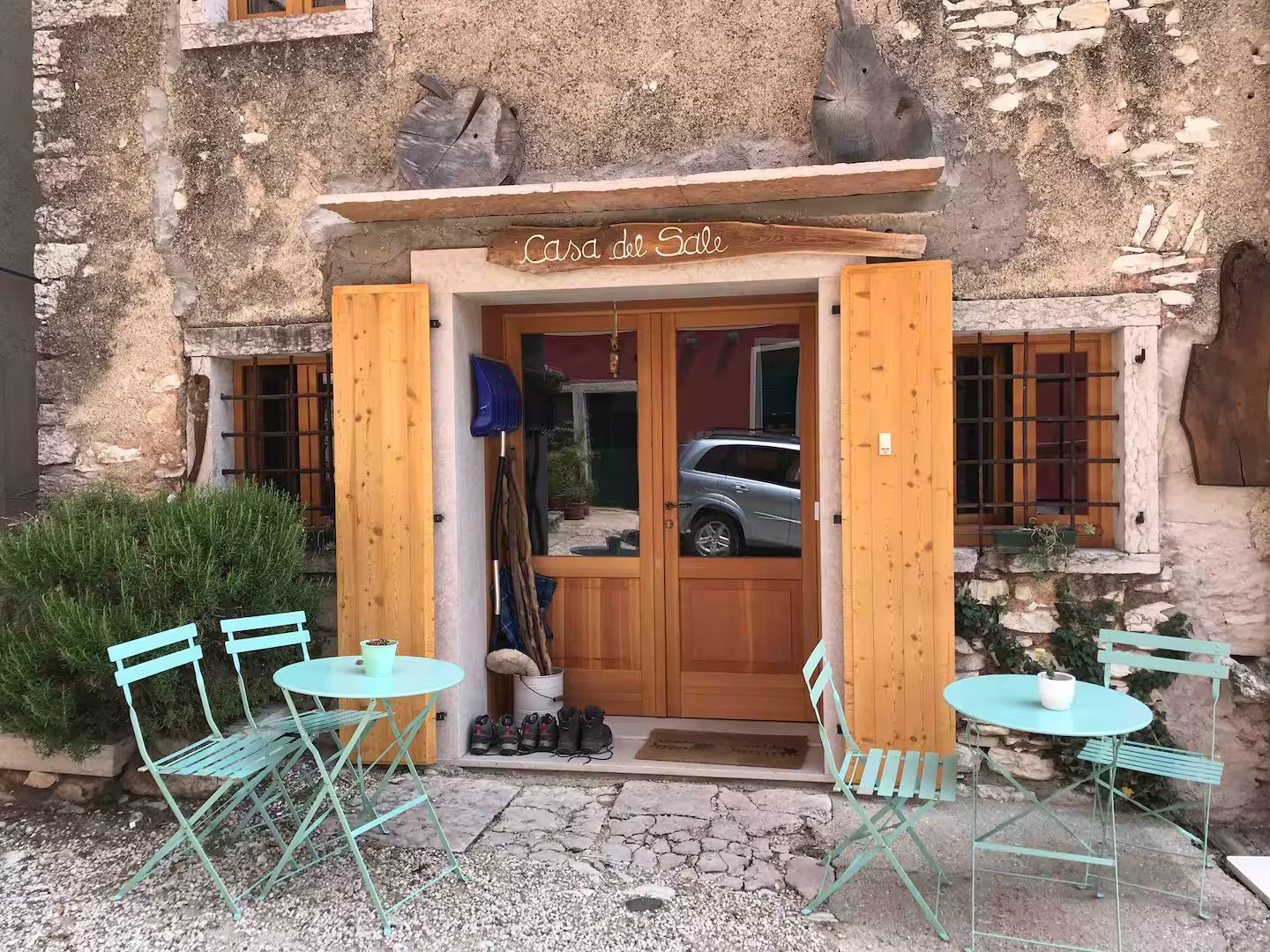 Esterno di un bar con porta e persiane in legno, tavoli e sedie color verde acqua. Edificio in pietra.