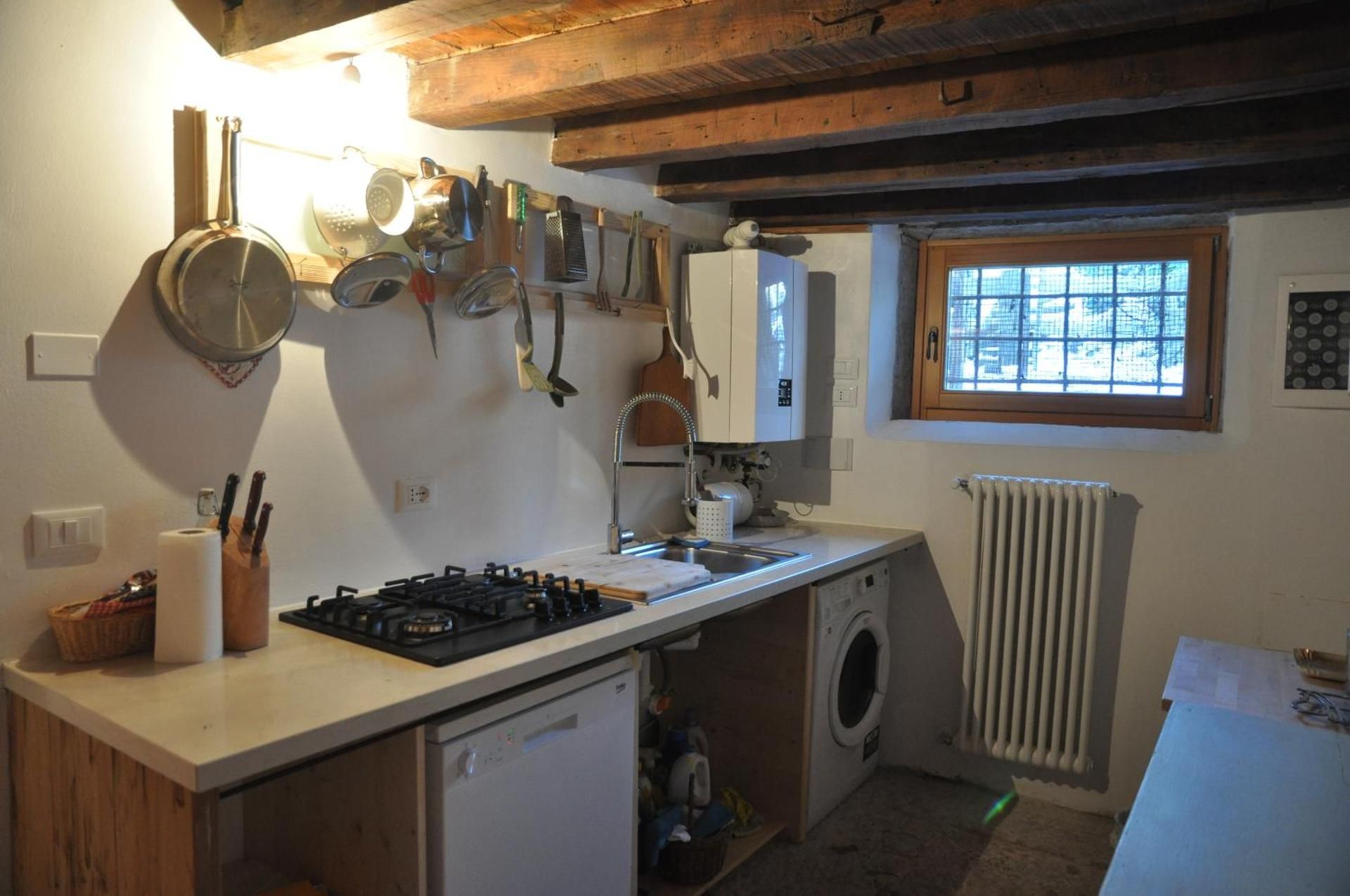 Piccola cucina rustica con elettrodomestici e pentole. Travi in legno, piccola finestra, pareti bianche.