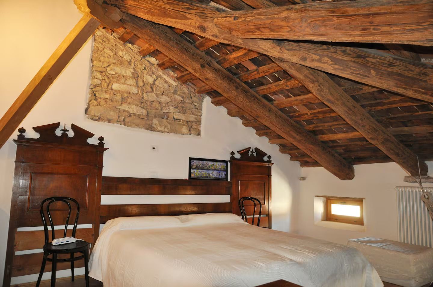 Camera da letto con letto, testiera in legno antico, travi in legno a vista e piccola finestra.