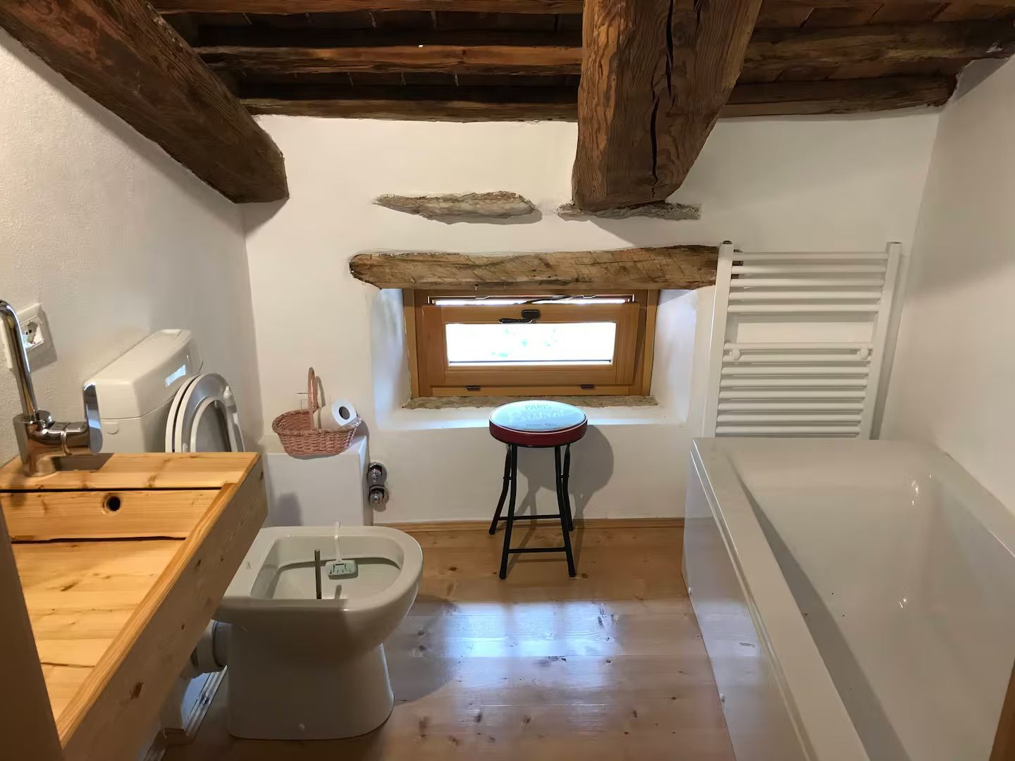 Bagno con travi in legno a vista, vasca da bagno, WC, lavandino e piccola finestra.