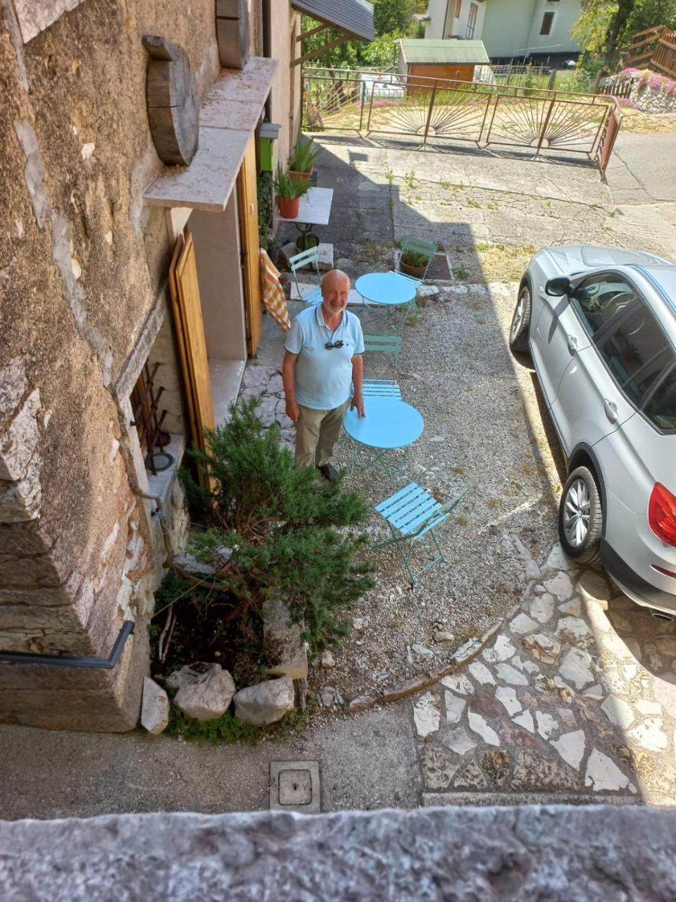 Un uomo in piedi in un cortile di pietra con sedie azzurre e un'auto argentata parcheggiata lì vicino.