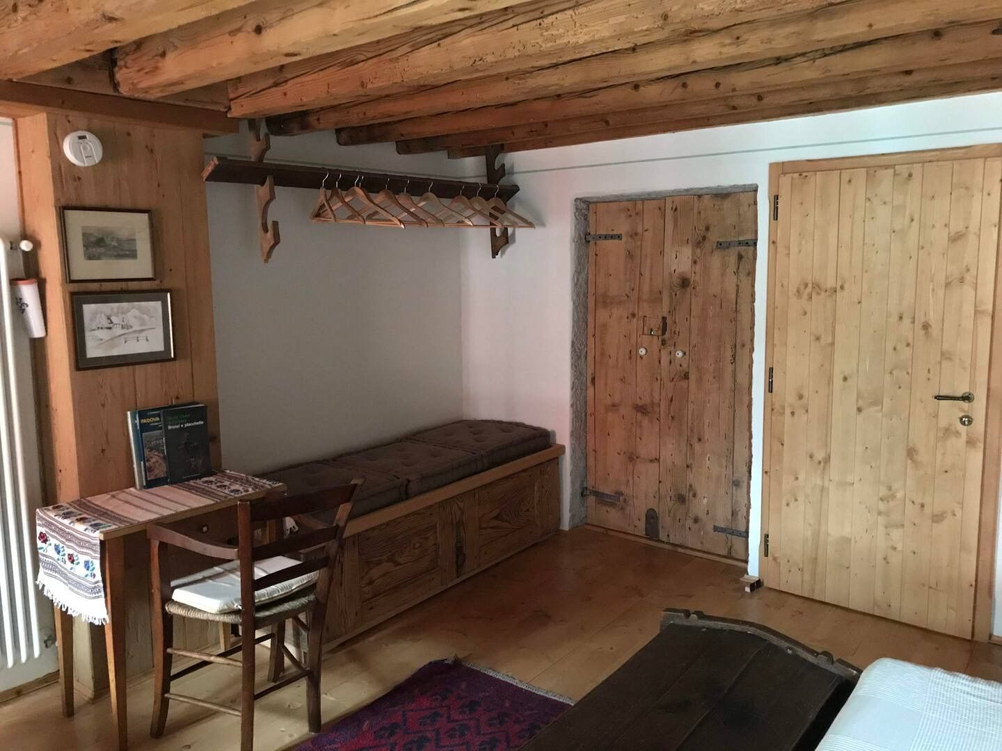 Stanza accogliente con travi in legno, panca contenitore, due porte in legno, un tavolino e appendiabiti.