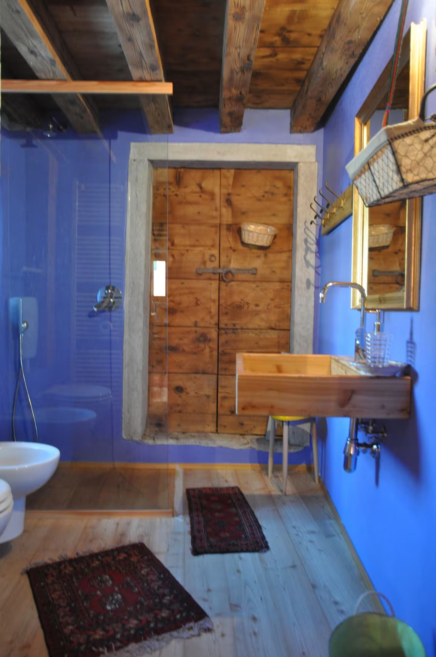 Bagno blu con porta, lavandino e travi in legno rustico. Tappeti sul pavimento in legno.