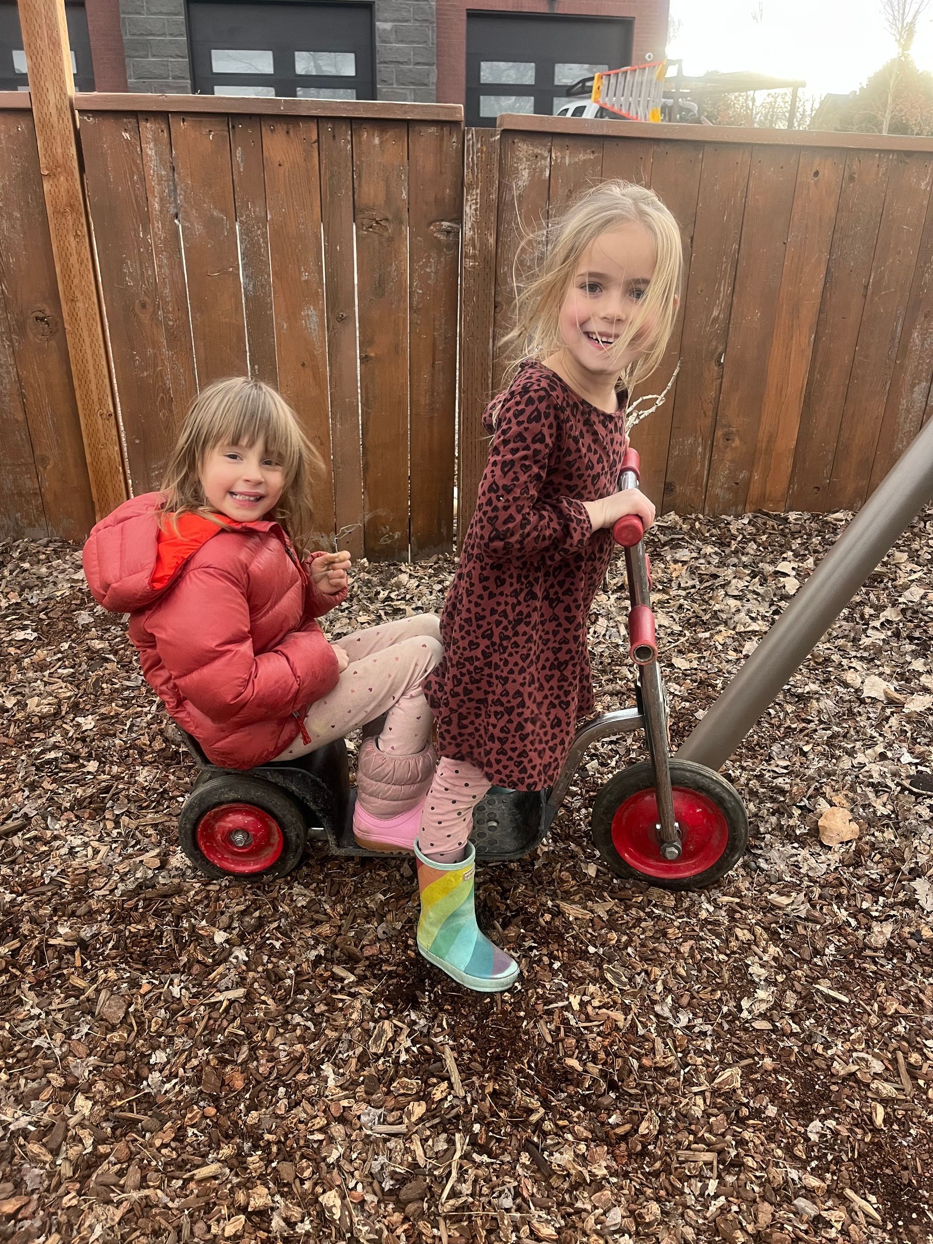 Two children ride a red scooter in a woodchip yard, smiling near a wooden fence.