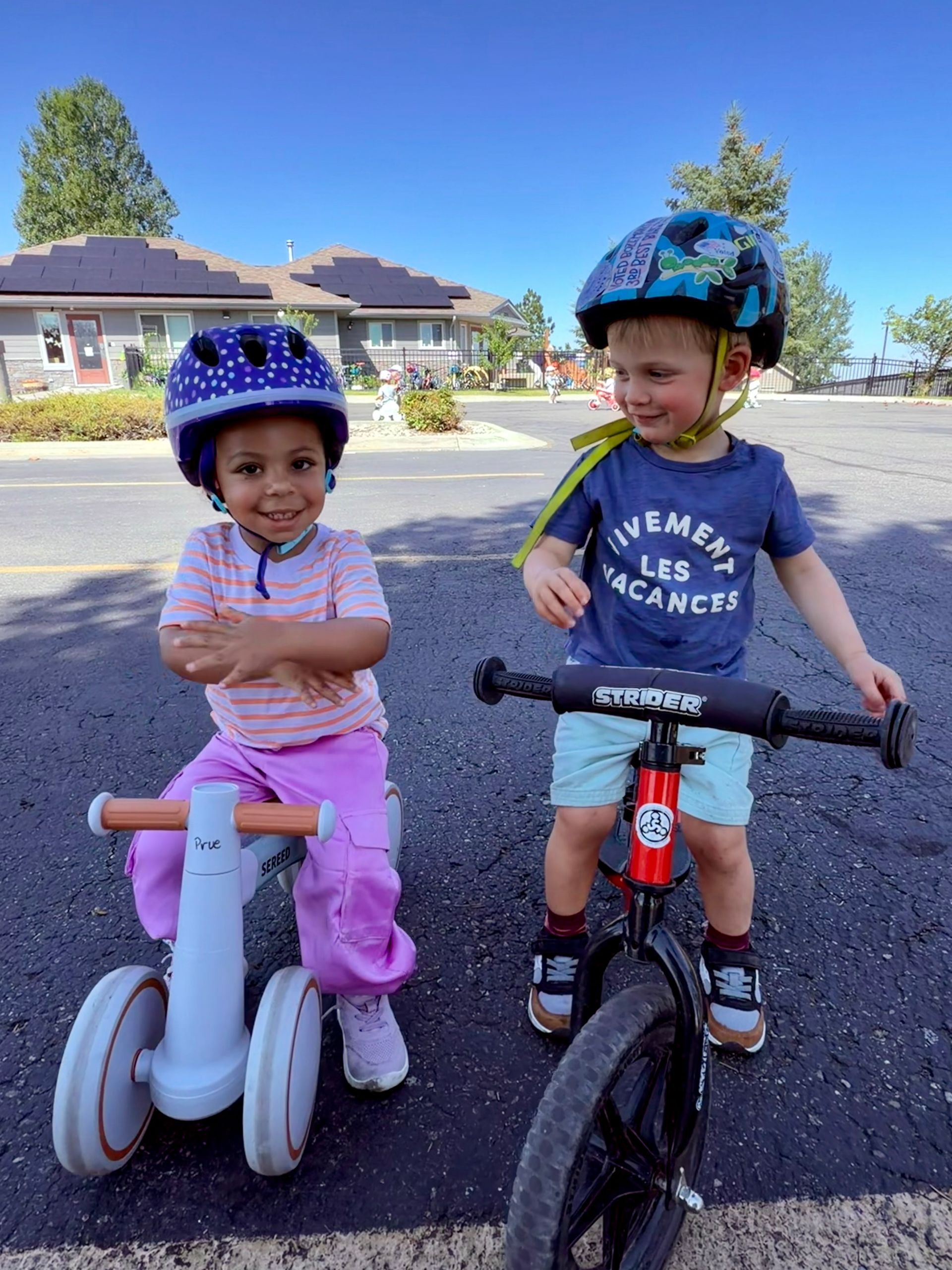 Two children wearing helmets ride balance bikes on a sunny neighborhood street.