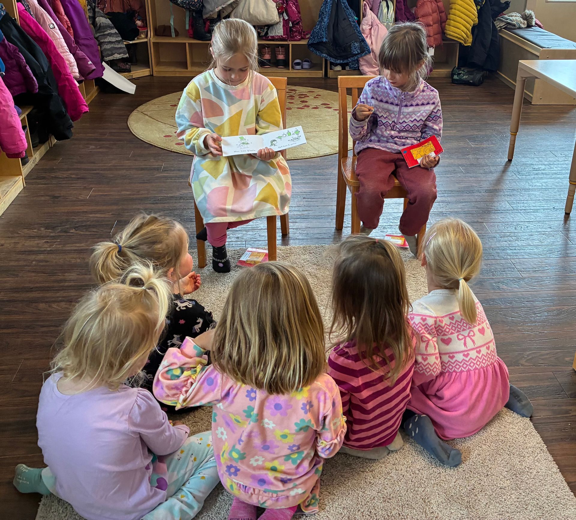 Children sitting on a rug listen to a child reading a book in a classroom.