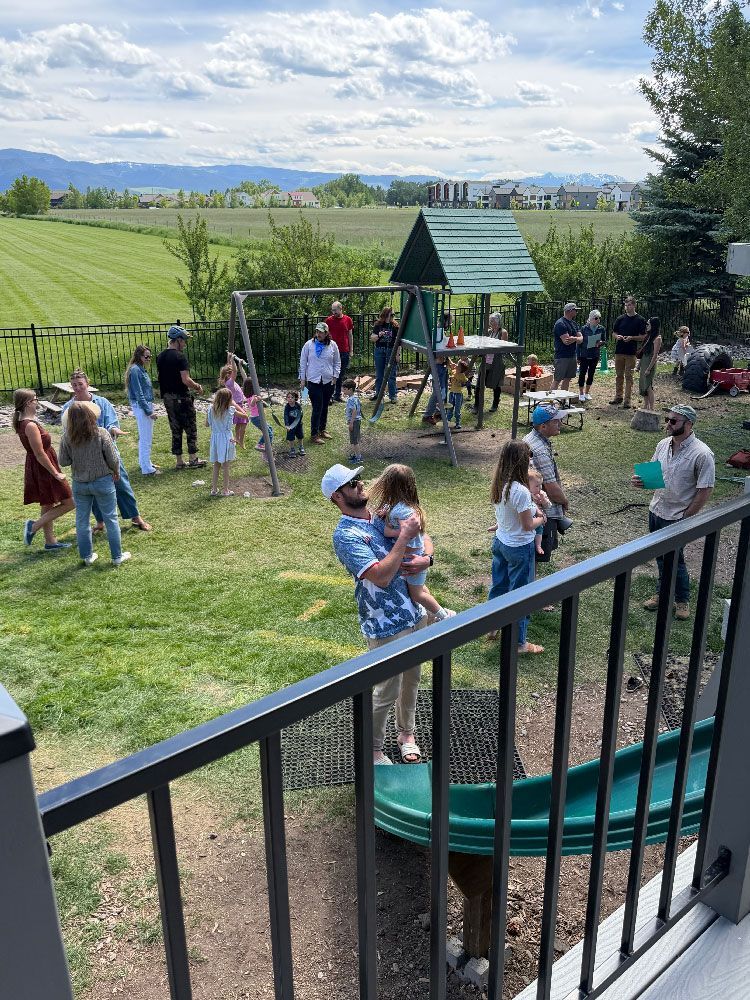 People gathered at a grassy playground on a sunny day, with mountains in the distance.