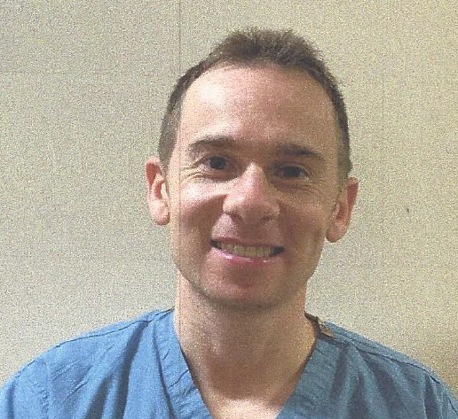 Man wearing blue scrubs smiles at the camera.