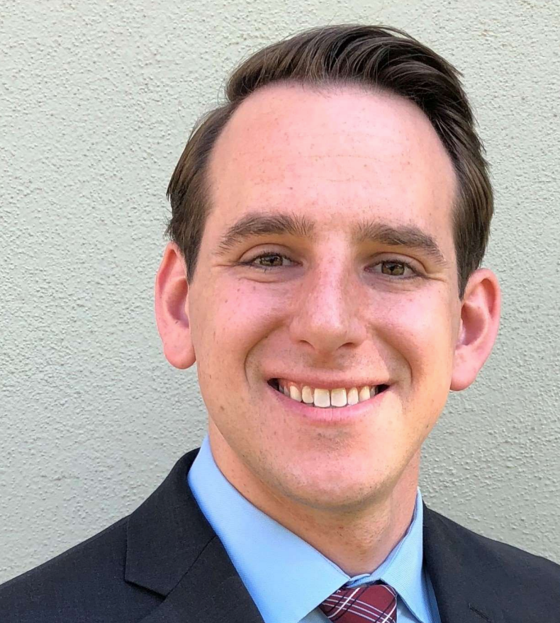 Man in suit smiling in front of a light wall. He has short brown hair and a patterned tie.