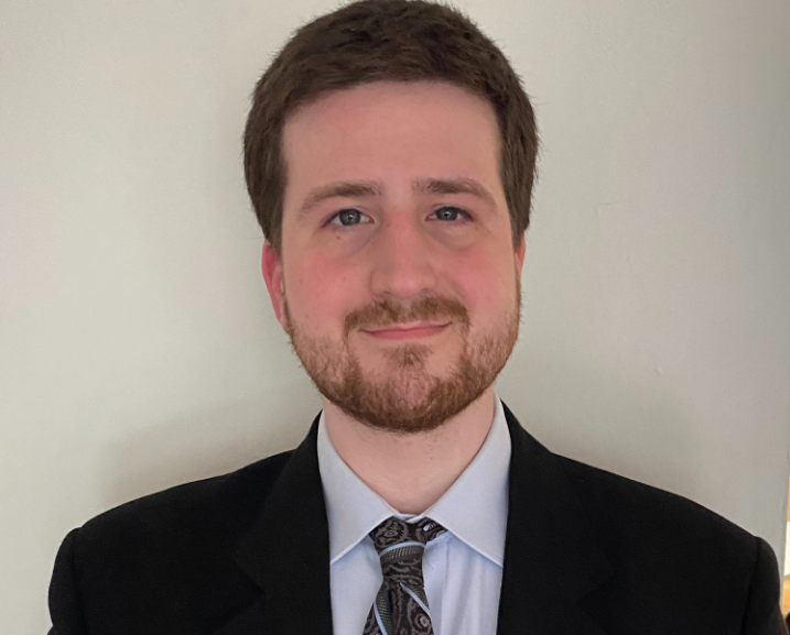Man with brown hair and beard, wearing a suit and tie, smiling at the camera.