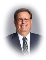 Man wearing glasses, suit, and tie smiling, against a blurred background.