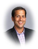 Smiling man in a dark blazer and patterned shirt against a light gray background.