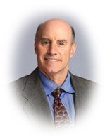 Man in suit smiles, looking at the viewer. He has a blue shirt and patterned tie, against a blurred background.