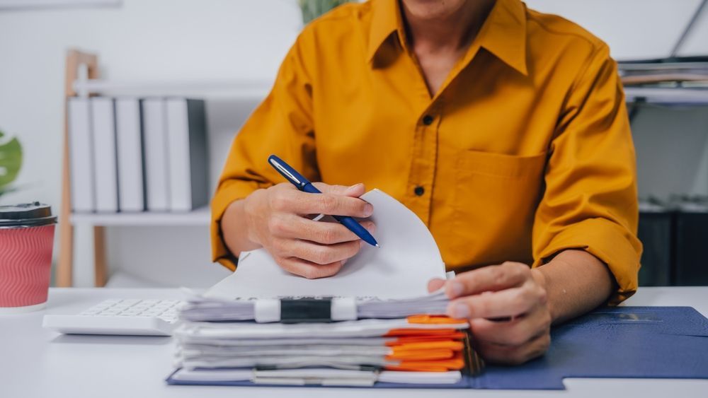 Person in a mustard yellow shirt writing in a notebook at a desk with stacked papers and folders