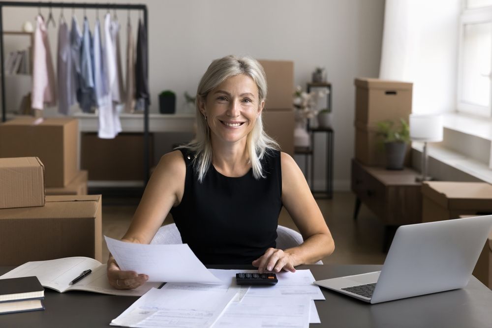 Woman at a desk in a moving office, reviewing papers beside a laptop and packed boxes.