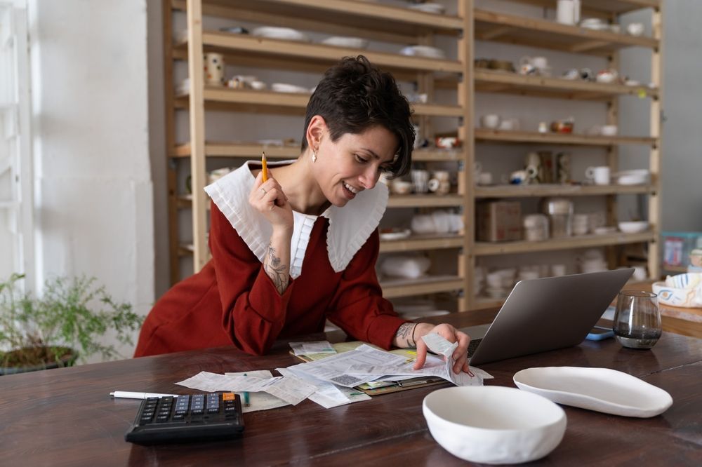 Person in red sweater sitting at a table, looking stressed while reviewing paperwork beside a laptop and calculator.
