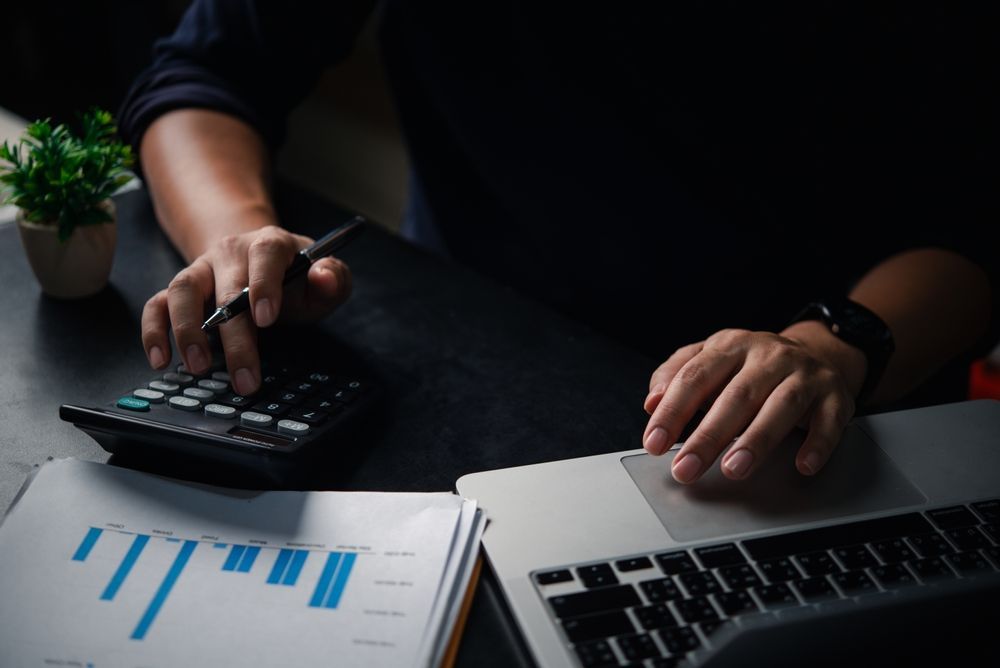 Hands using calculator and laptop beside a paper chart on a dark desk